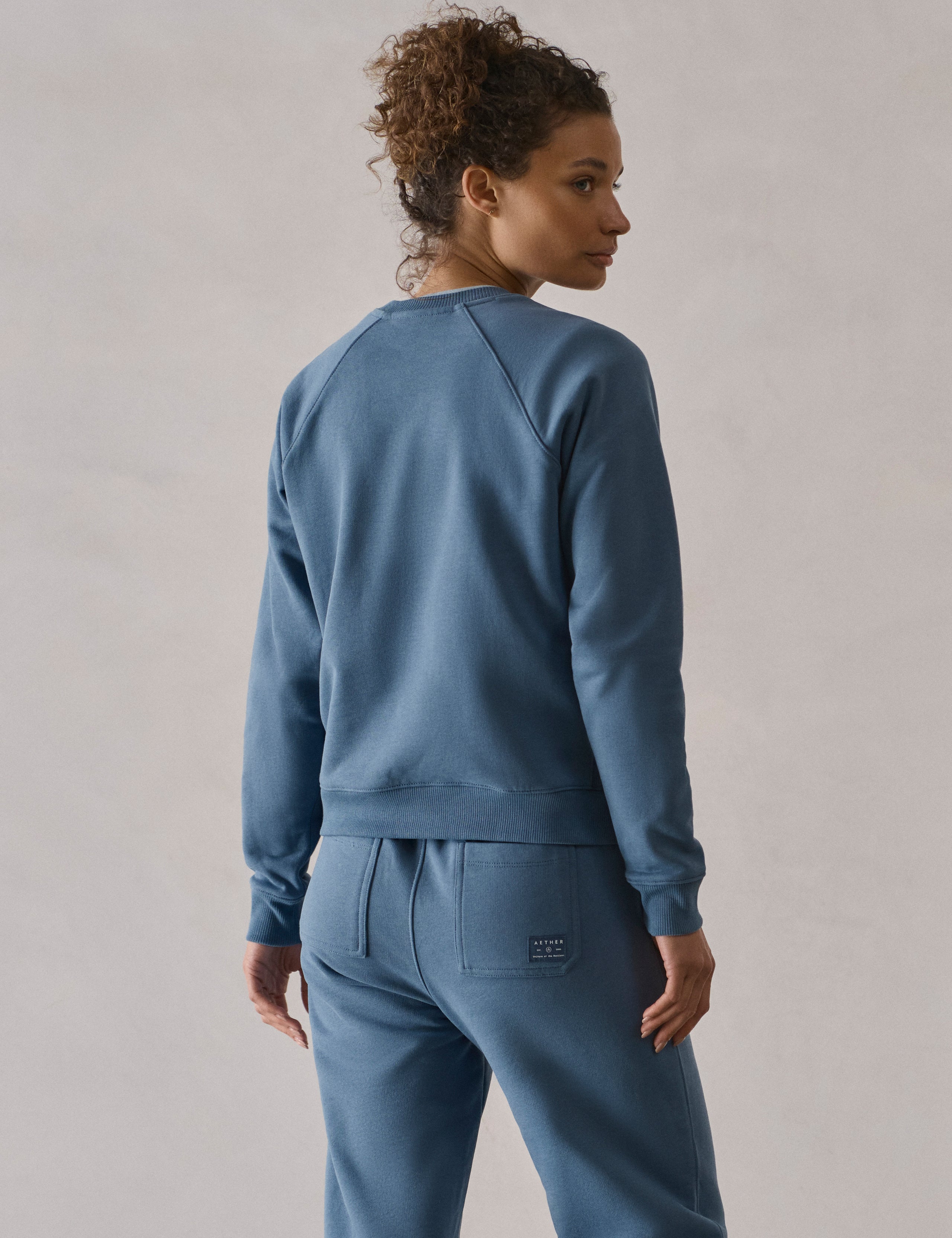 Rear 3/4 upper-body view of woman wearing W Osa Raglan Sweatshirt in Coastal Blue from AETHER Apparel.