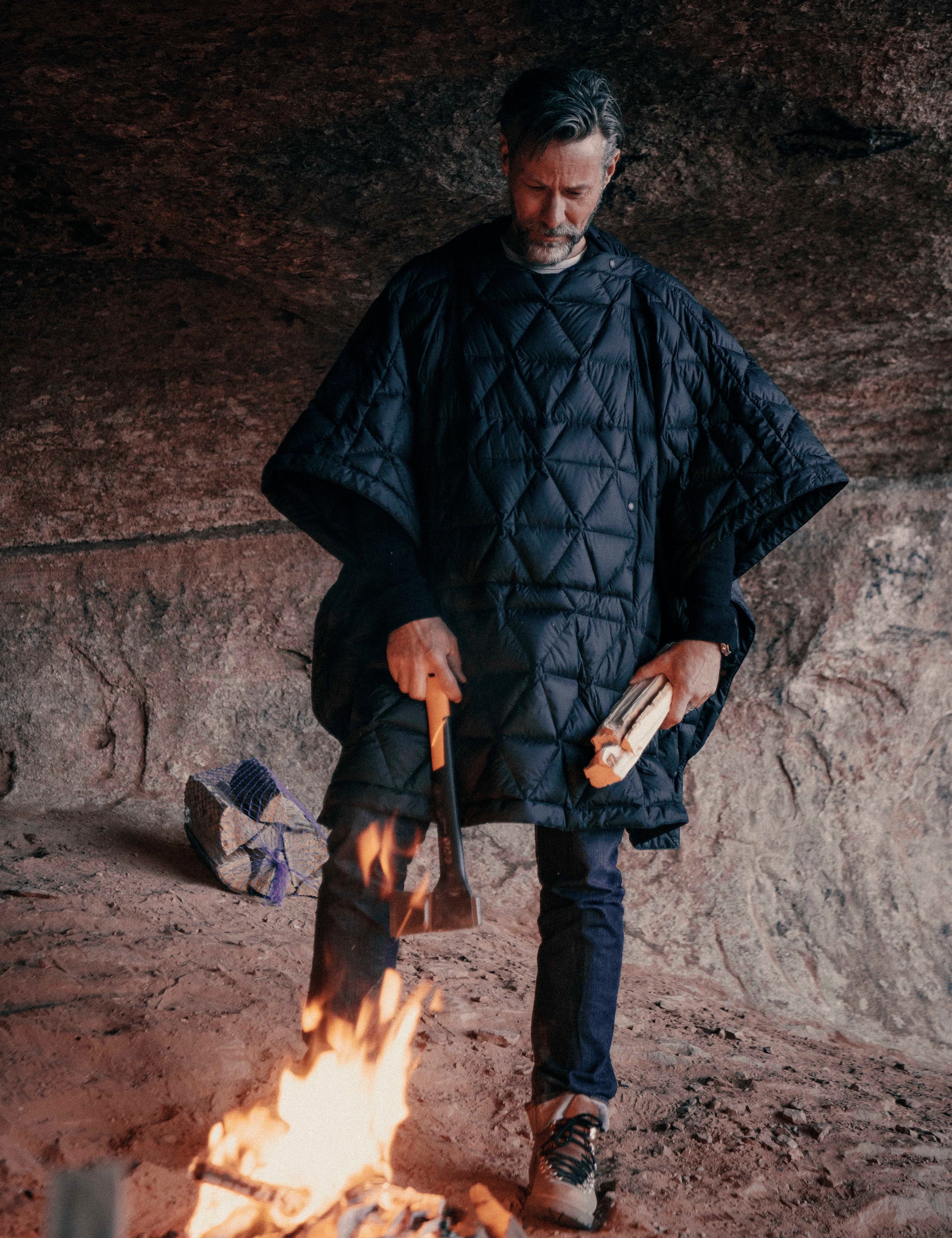 Outdoor lifestyle image of man standing over campfire wearing Down Poncho in Onyx Black from AETHER Apparel.