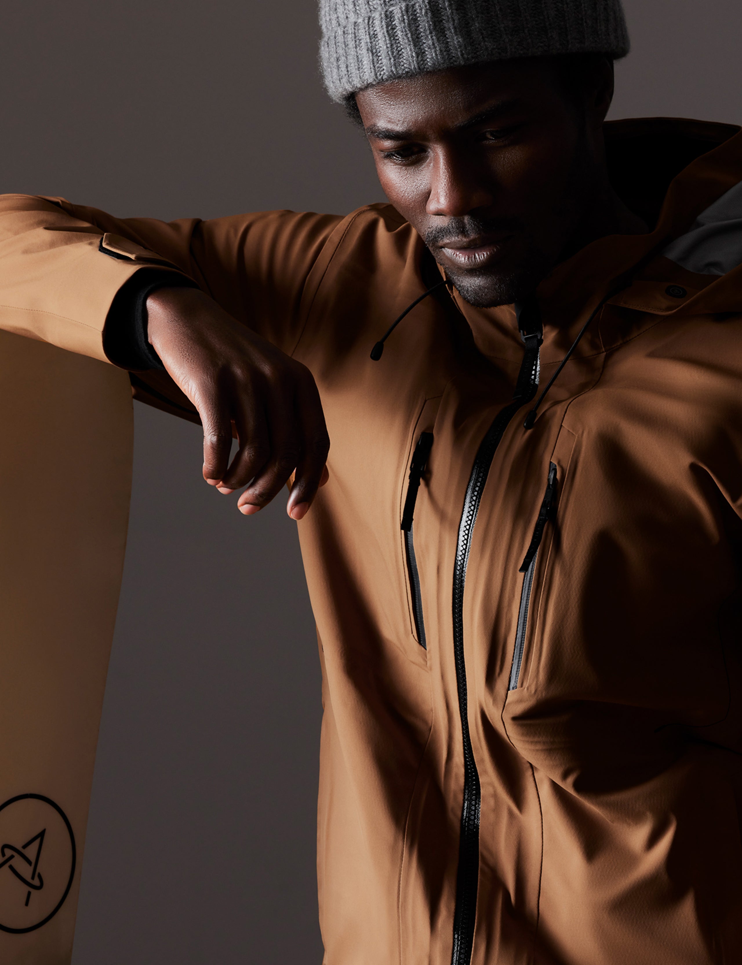 Closeup view of man leaning on snowboard wearing Altitude Snow Shell 2.0 in Rubber Brown from AETHER Apparel.