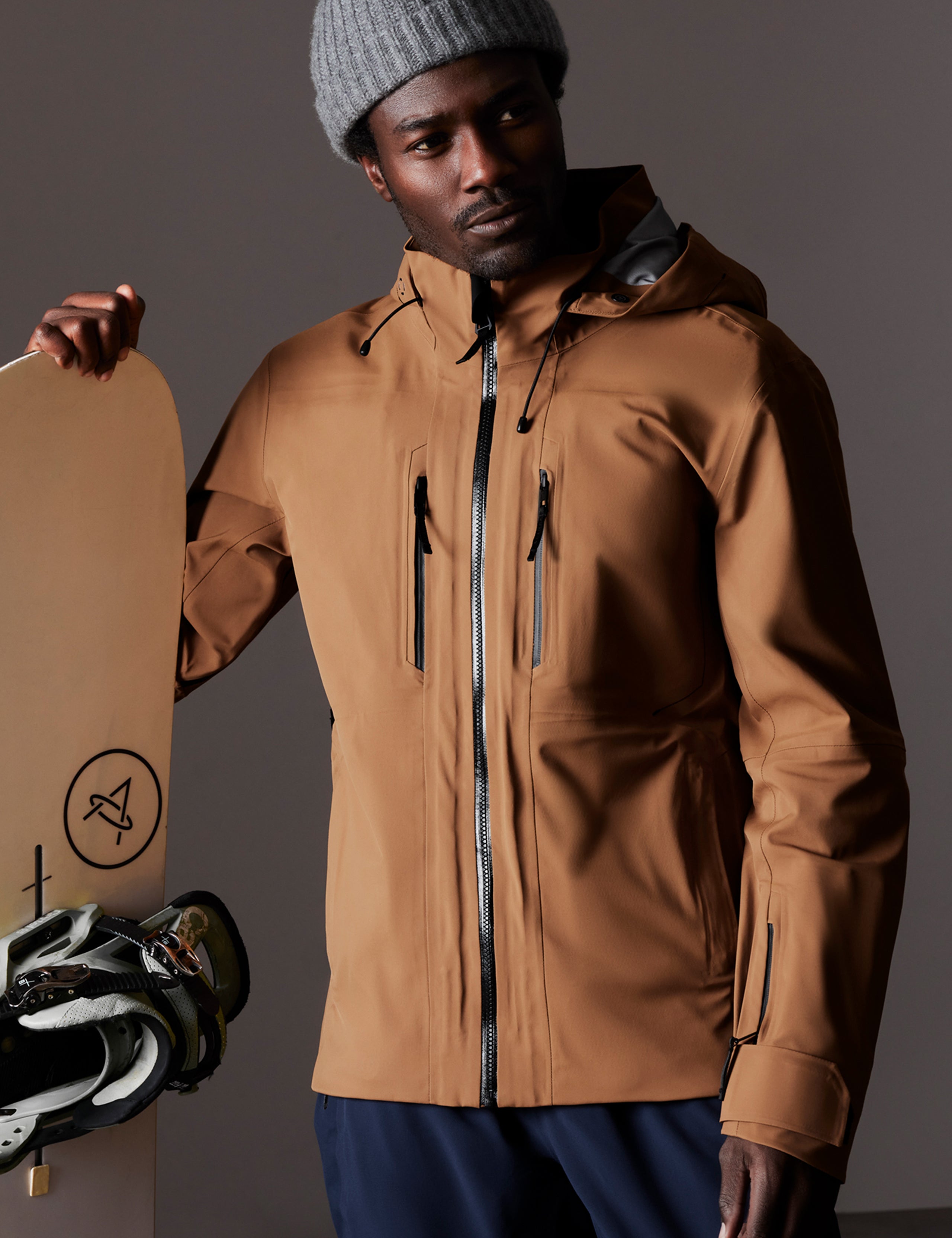 Front body view of man holding snowboard wearing Altitude Snow Shell 2.0 in Rubber Brown from AETHER Apparel.