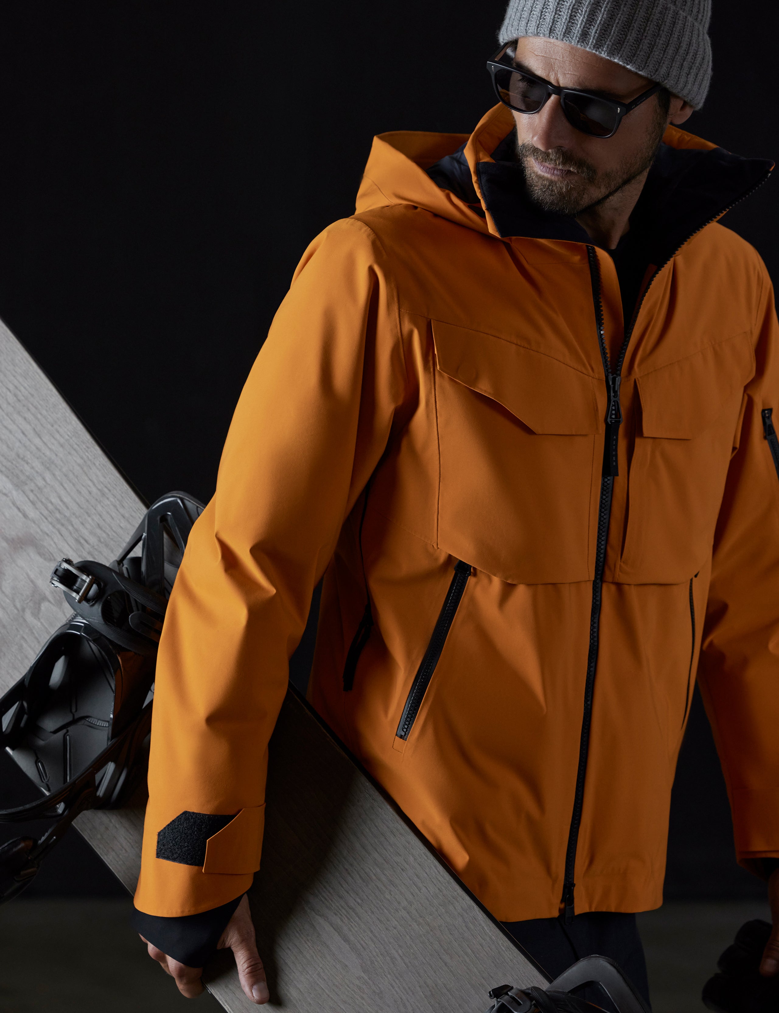 Front 3/4 body view of man holding snowboard wearing Brighton Insulated Snow Jacket in Autumn Orange from AETHER Apparel.