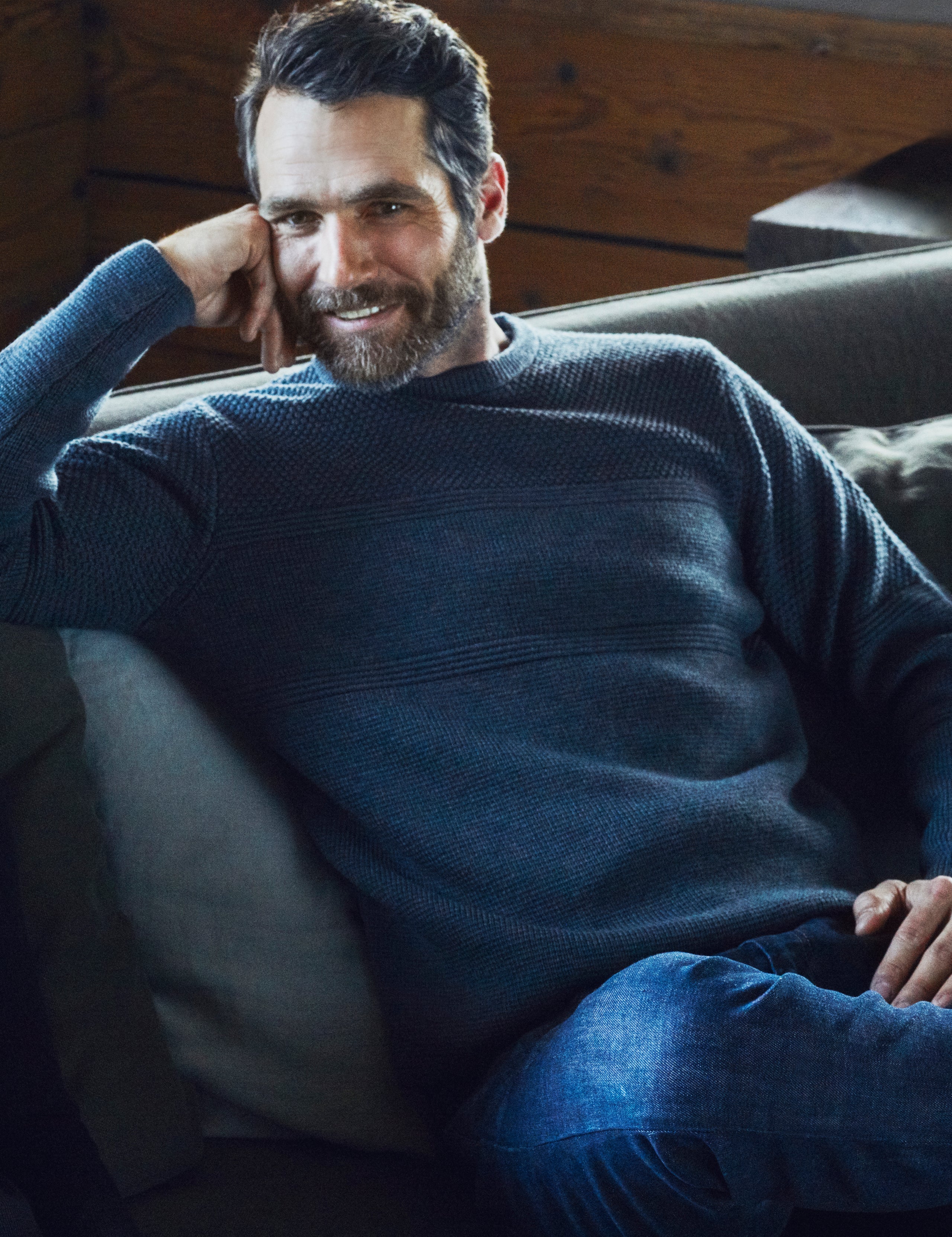 Lifestyle image of man sitting on couch wearing Cooper Sweater in Dark Charcoal Heather from AETHER Apparel.