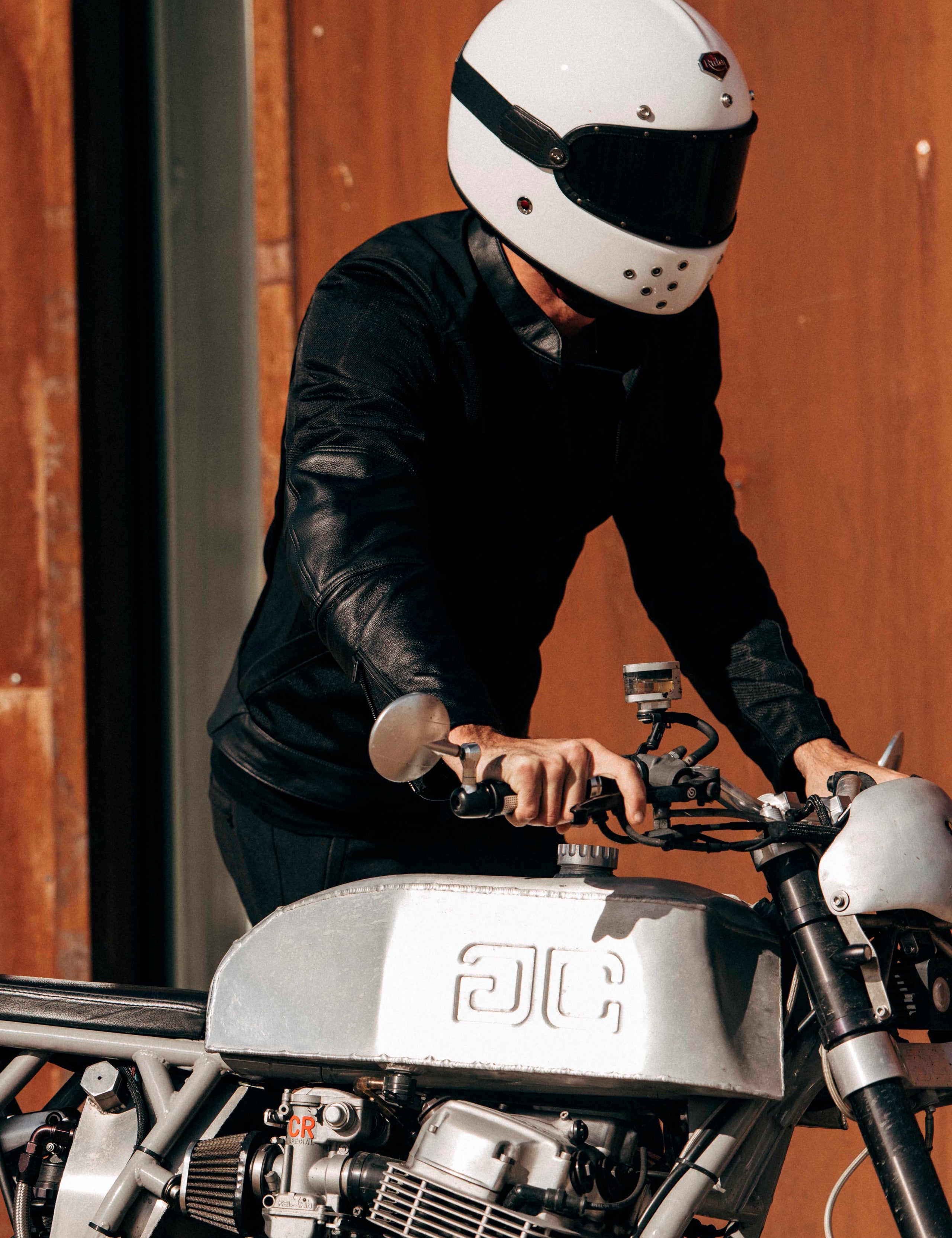 Outdoor lifestyle image of man pushing custom motorcycle wearing white helmet and Draft Mesh Motorcycle Jacket in Jet Black from AETHER Apparel.