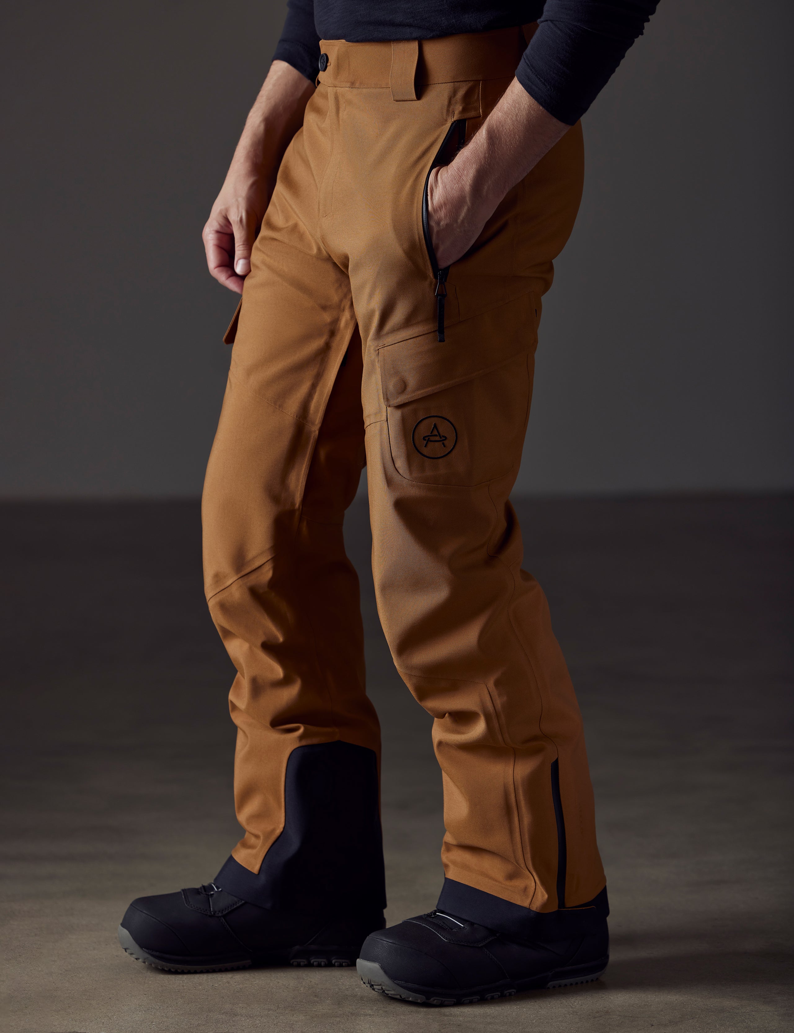 Front angled lower body view of man with knees bent and hand in pant pocket wearing Explorer Snow Pant in Rubber Brown from AETHER Apparel.