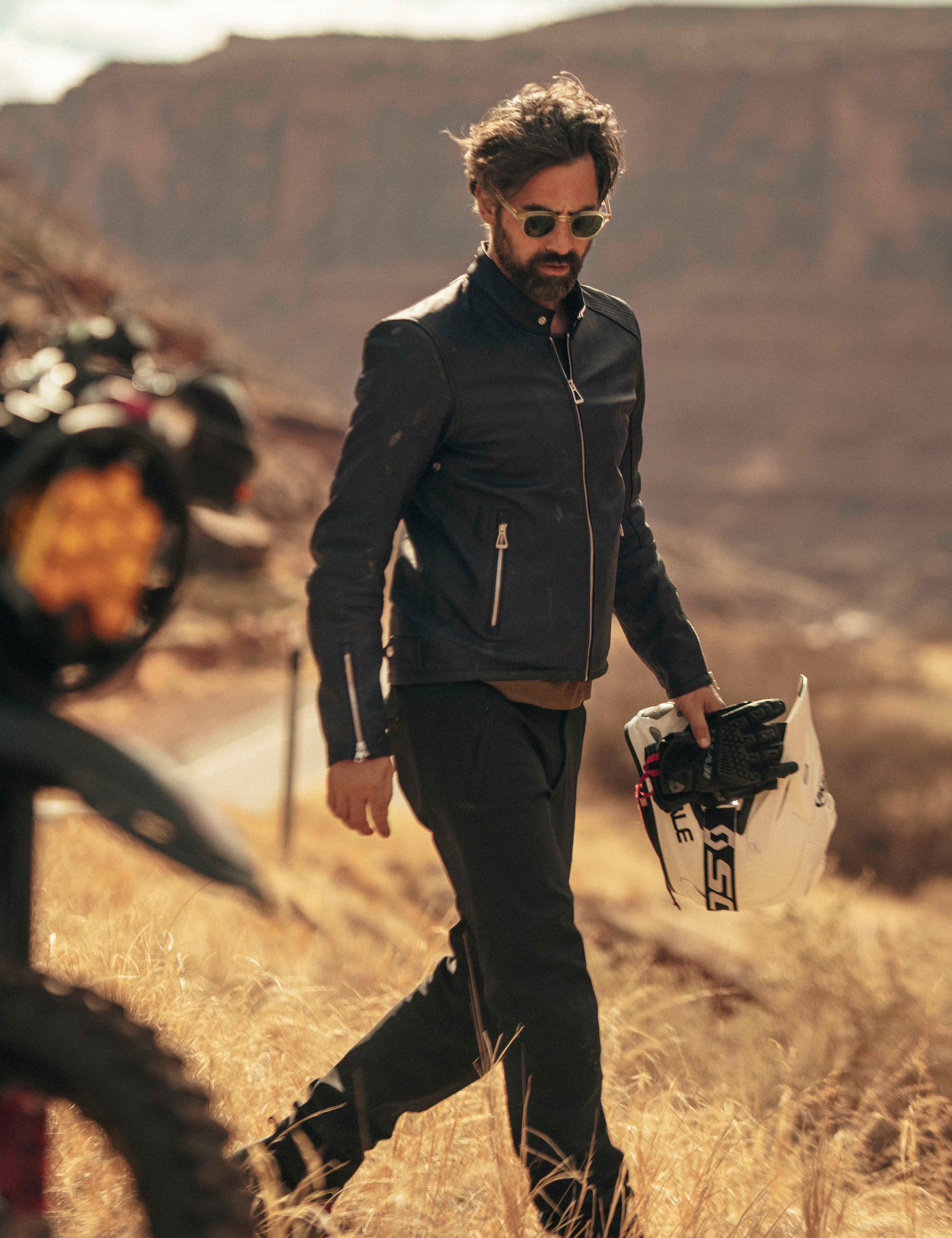 Outdoor lifestyle image of man walking past motorcycle wearing Laslo Motorcycle Jacket in Onyx Black from AETHER Apparel.