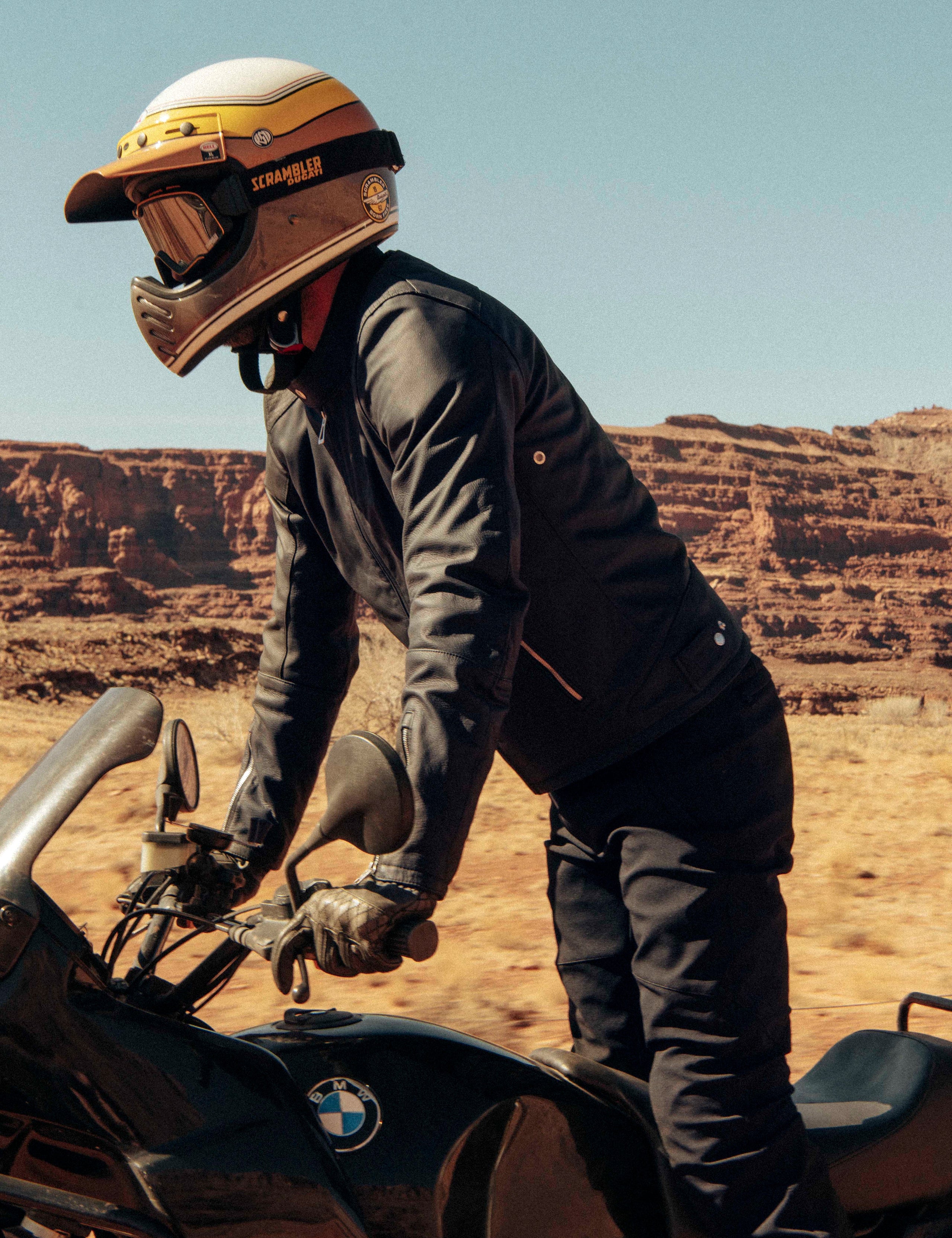 Outdoor lifestyle image of man standing up on BMW motorcycle wearing Laslo Motorcycle Jacket in Onyx Black from AETHER Apparel.