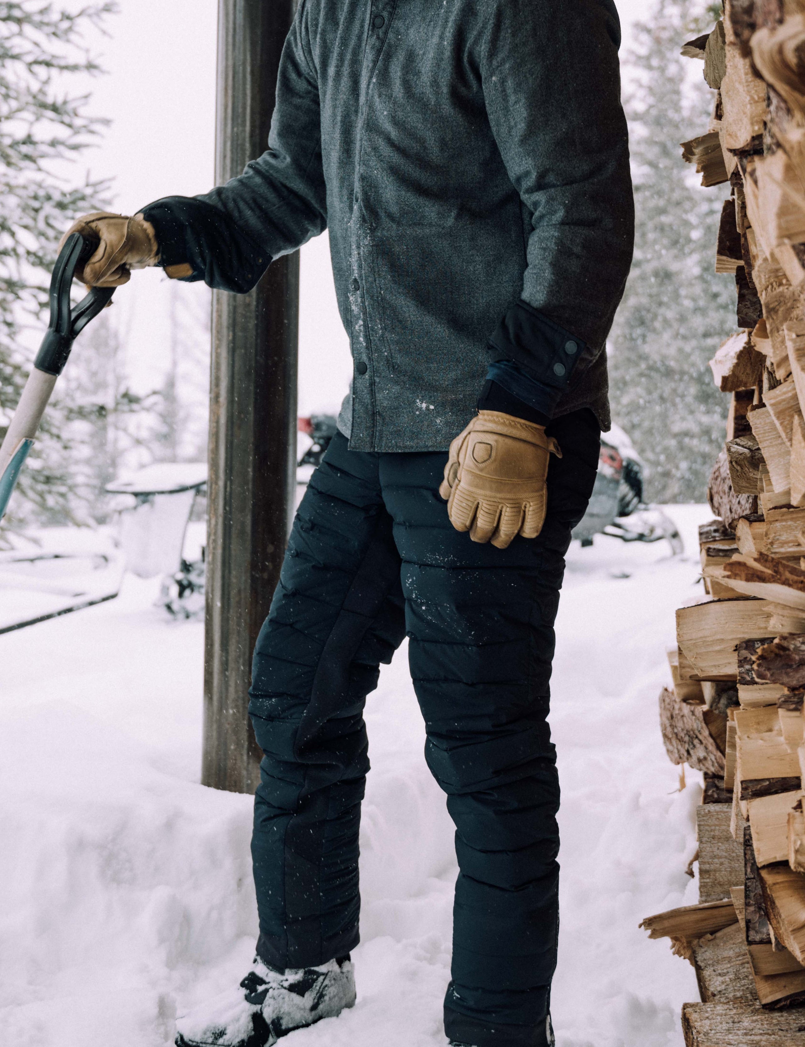 Outdoor lifestyle image of man holding shovel next to firewood pile wearing Launch Pant in Onyx Black from AETHER Apparel. 
