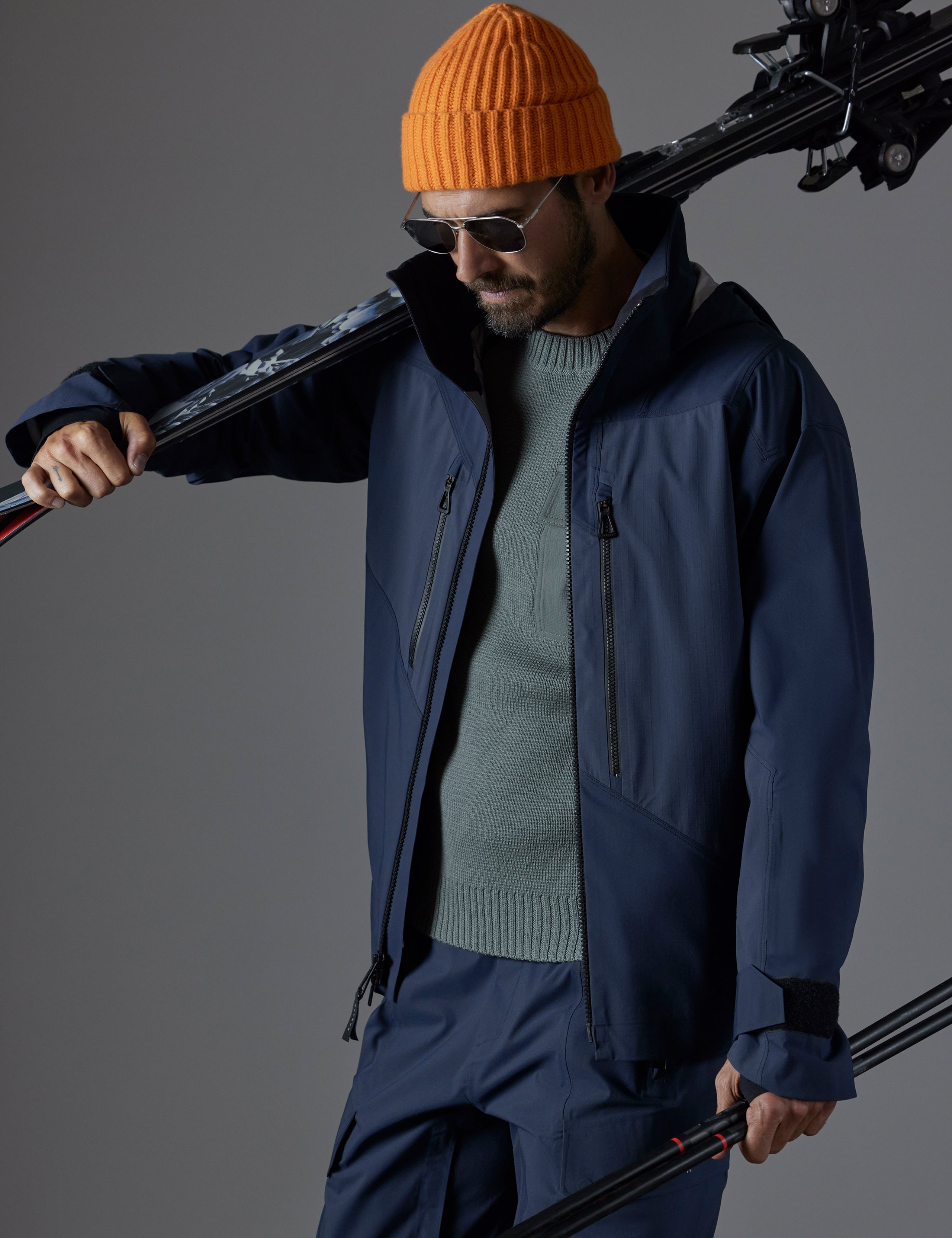 Front 3/4 full-body view of man holding skis over shoulder wearing Maverick Snow-Shell Jacket in Total Eclipse dark blue from AETHER Apparel.