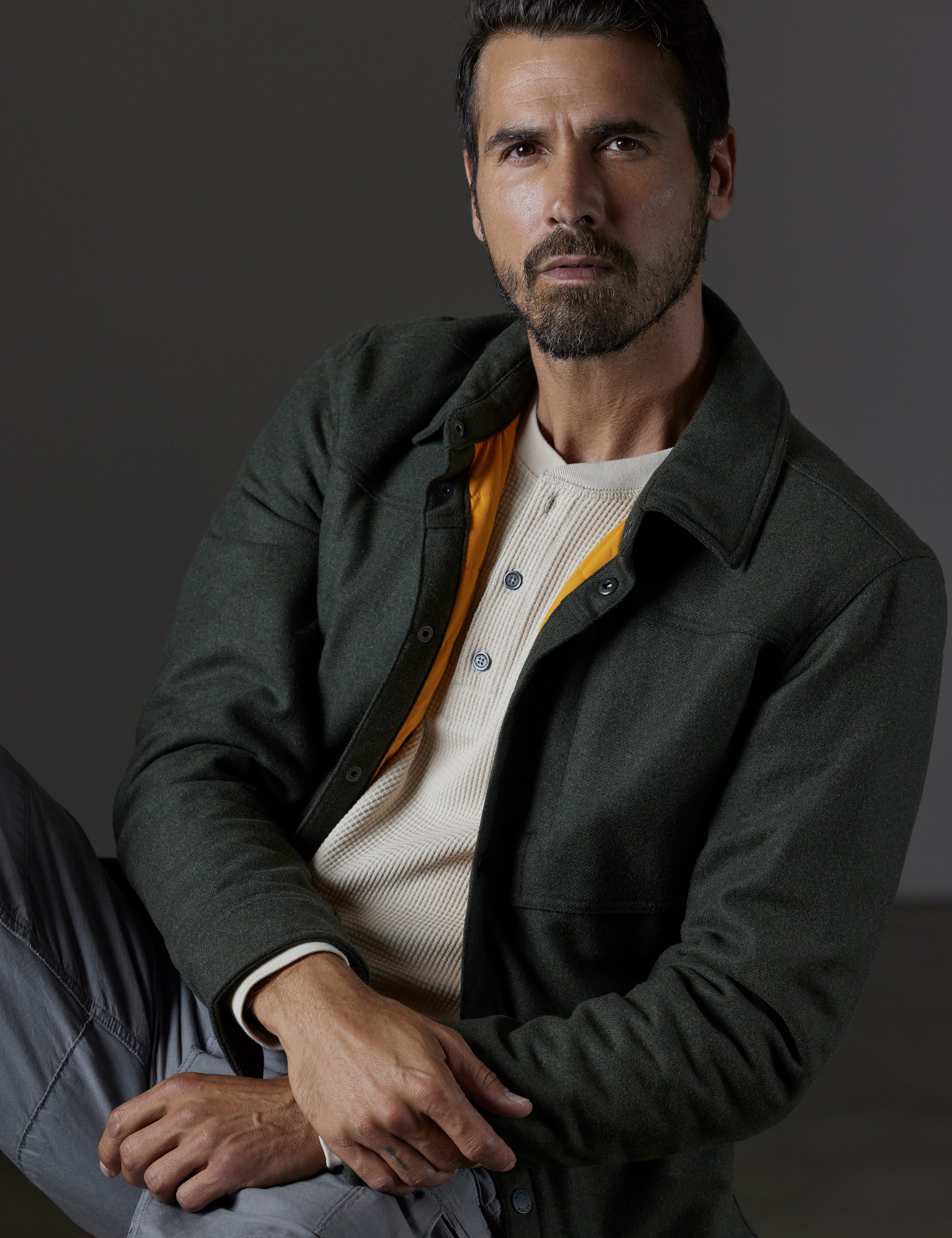 Front 3/4 body view of man sitting wearing unbuttoned Nova Insulated Button-Down in Command Green from AETHER Apparel.