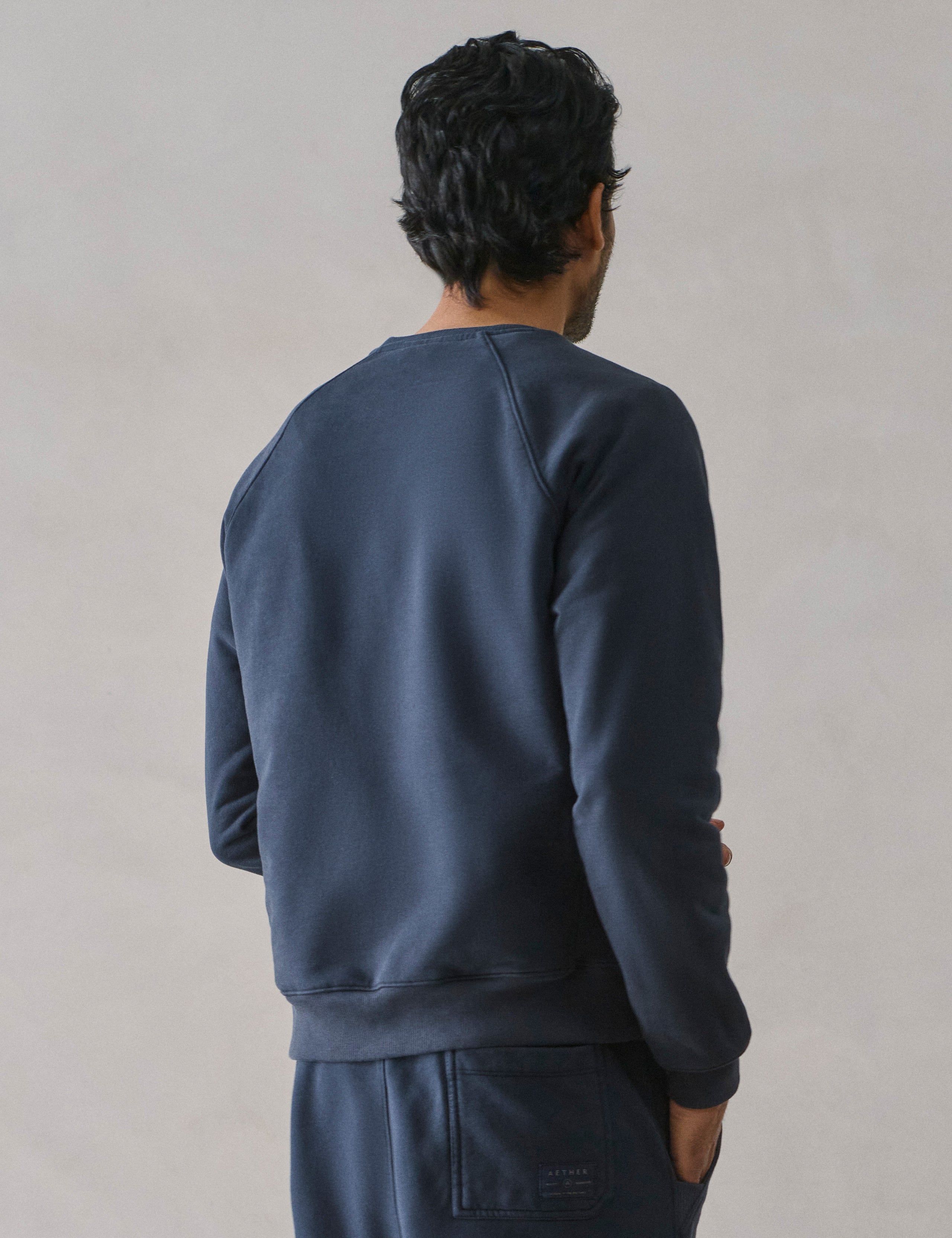 Rear 3/4 upper body view of man wearing Osa Raglan Sweatshirt in Total Eclipse dark blue from AETHER Apparel.