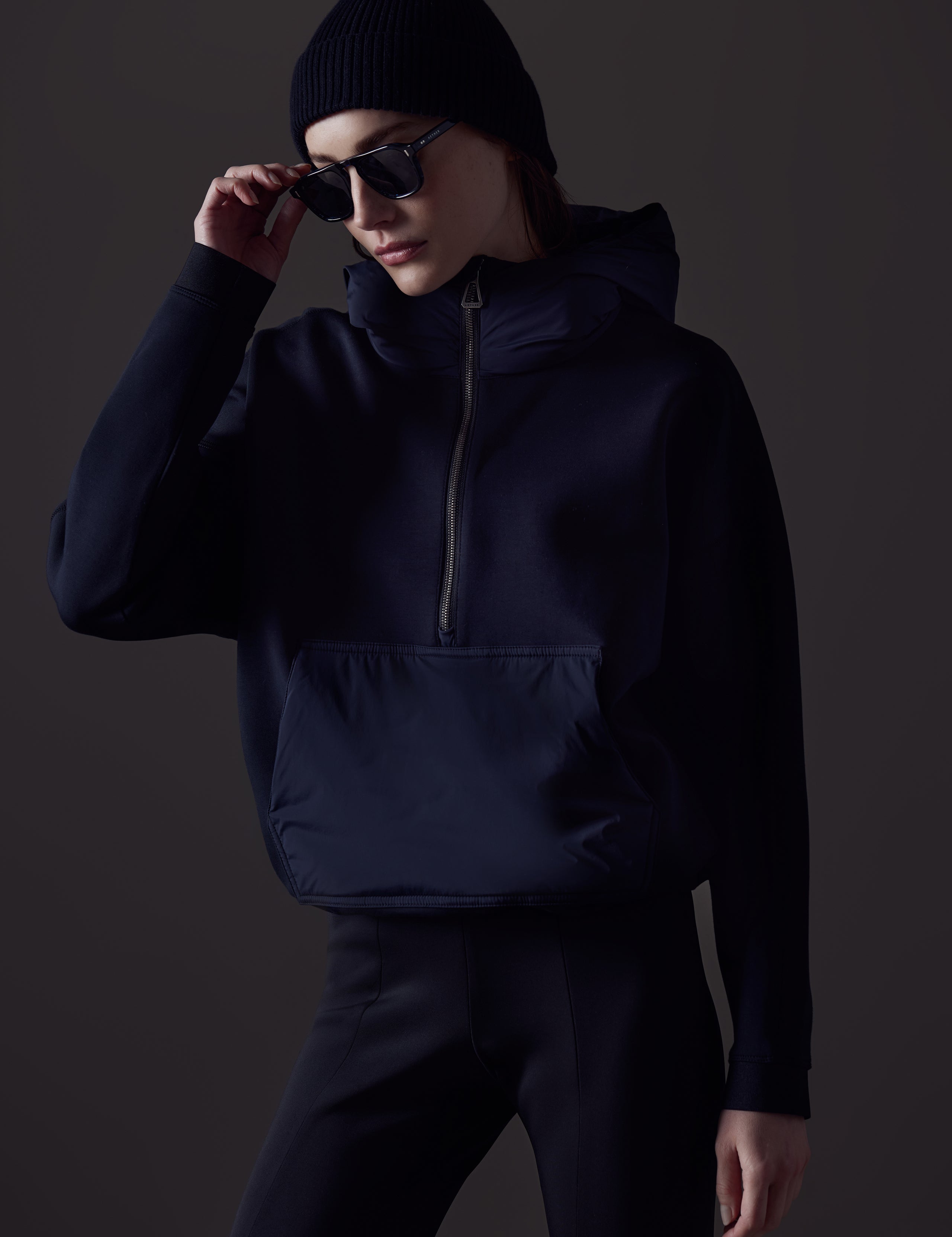 Front angled view of woman with sunglasses and beanie wearing Align Hooded Anorak in Onyx Black from AETHER Apparel.