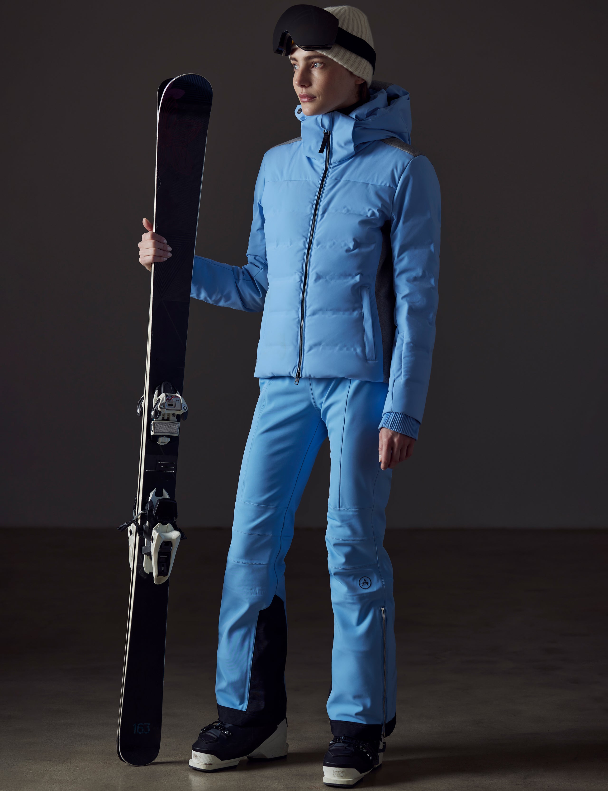 Front angled full-body view of woman holding skis wearing Bancroft Snow Jacket in Atlas Blue from AETHER Apparel.