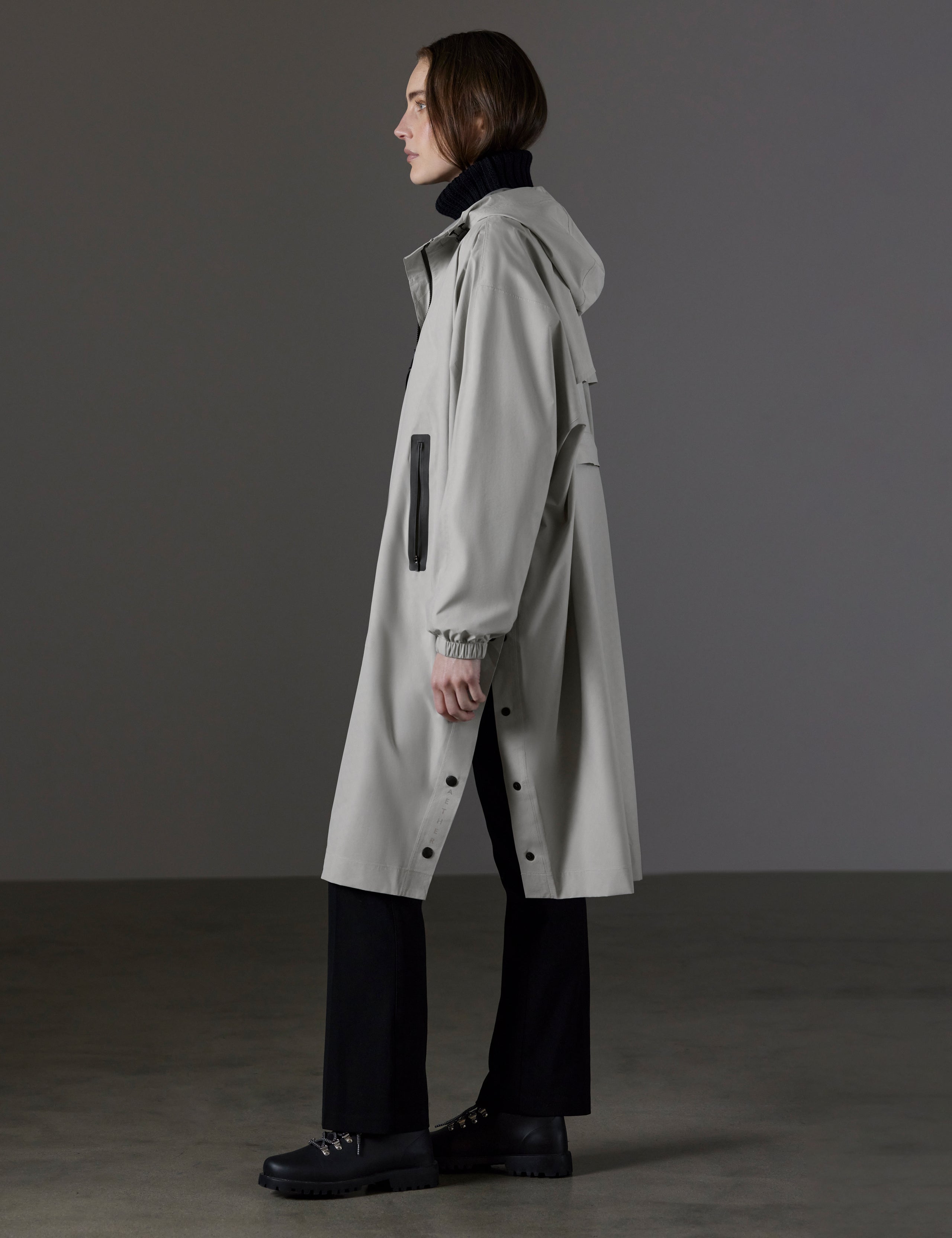 Side profile full-body view of woman wearing Kite Packable Rain Poncho in Stone Grey from AETHER Apparel.