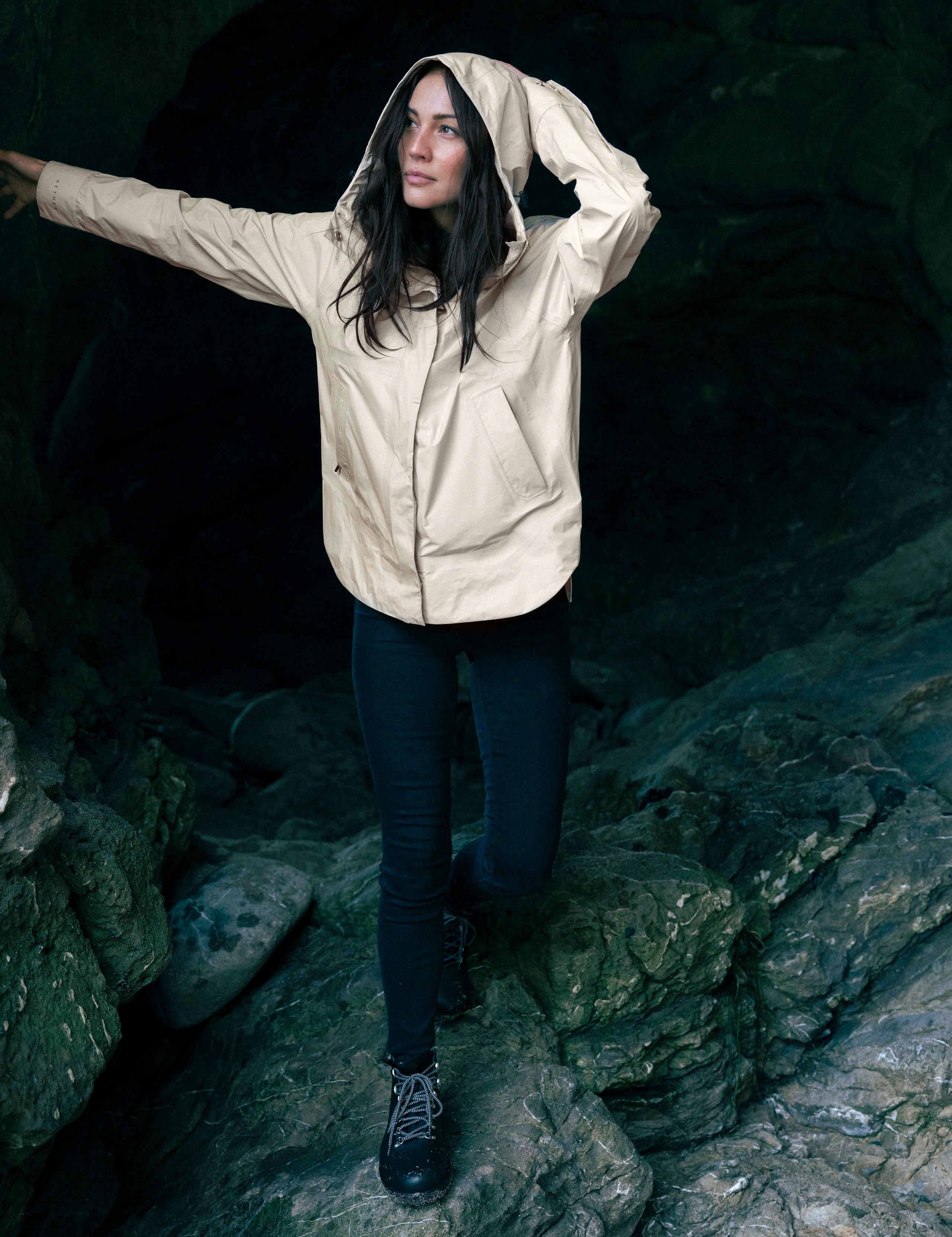 Outdoor lifestyle image of woman standing on rocks in cave wearing Storm All-Weather Jacket in Bone white from AETHER Apparel.